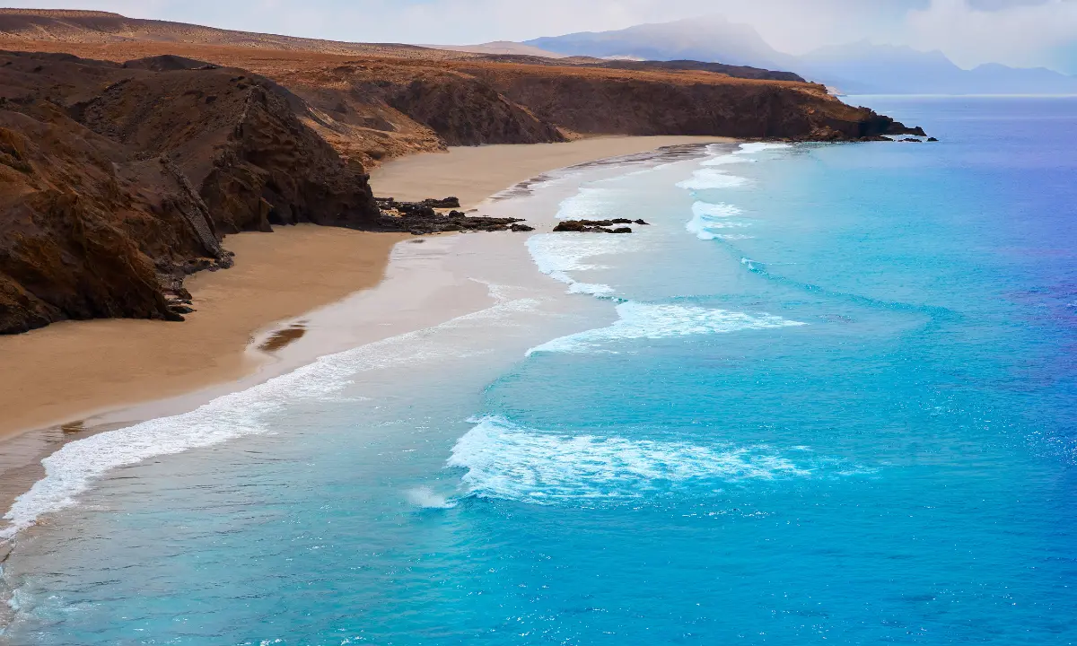 Playa de arena entre acantilados con olas azules y espuma blanca al atardecer