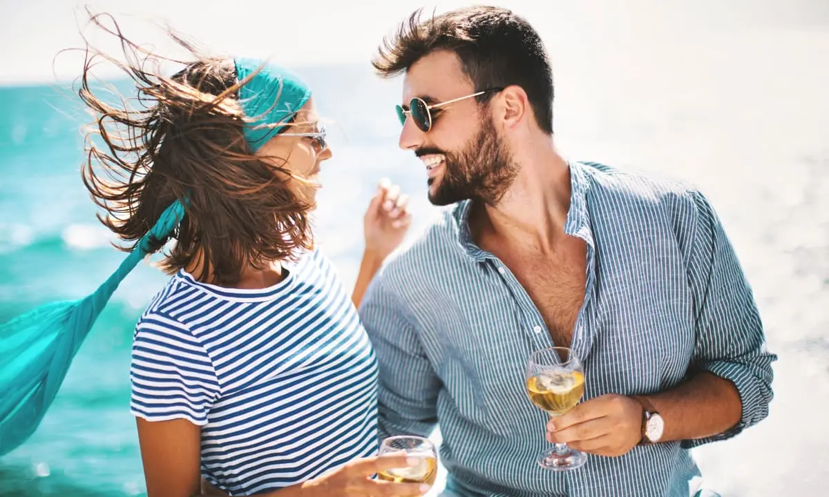 Pareja sonriente en la playa, con gafas y vasos de bebida al atardecer