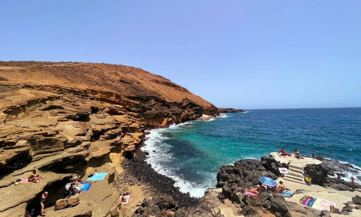 Playa rocosa con ba&ntilde;istas y el mar azul bajo un cielo despejado.