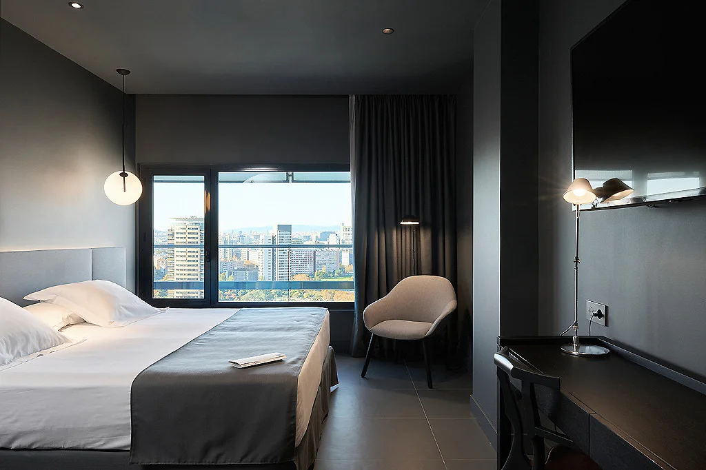 Dormitorio moderno con cama, cortinas oscuras, silla y vista urbana por ventana