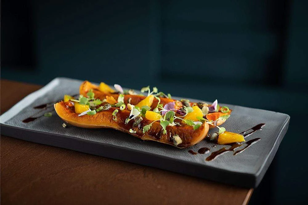 Tostada con calabaza, semillas y brotes sobre bandeja negra, con salsa roja.