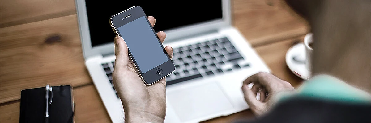 Persona sostiene un tel&eacute;fono mientras usa una computadora port&aacute;til en escritorio.