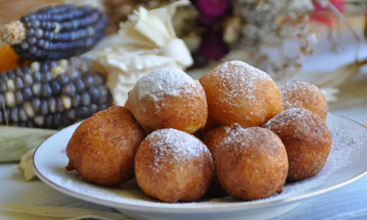 Bu&ntilde;uelos caseros dorados con az&uacute;car glass en un plato blanco sobre mesa.