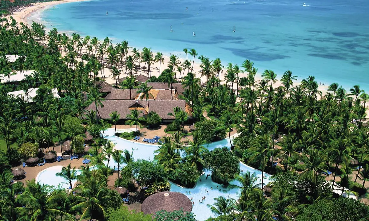 Vista a&eacute;rea de resort tropical con palmeras, piscina y playa de aguas turquesa