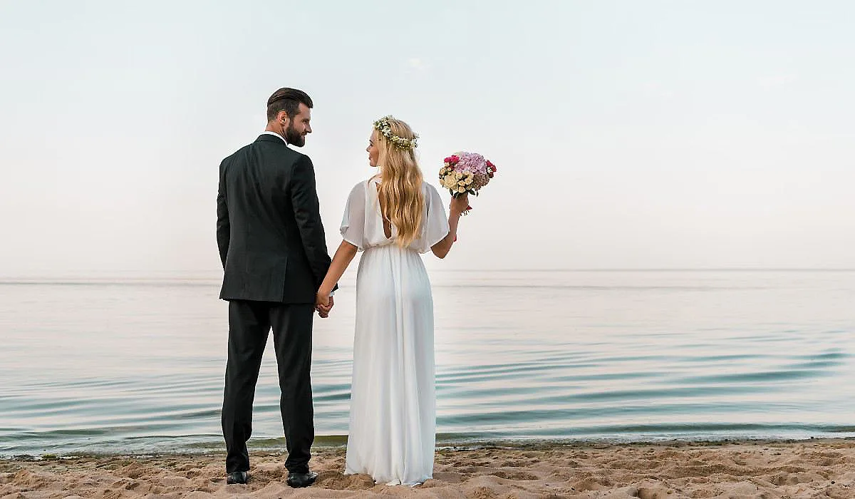 Pareja con vestido de novia y traje negro, caminando junto al mar