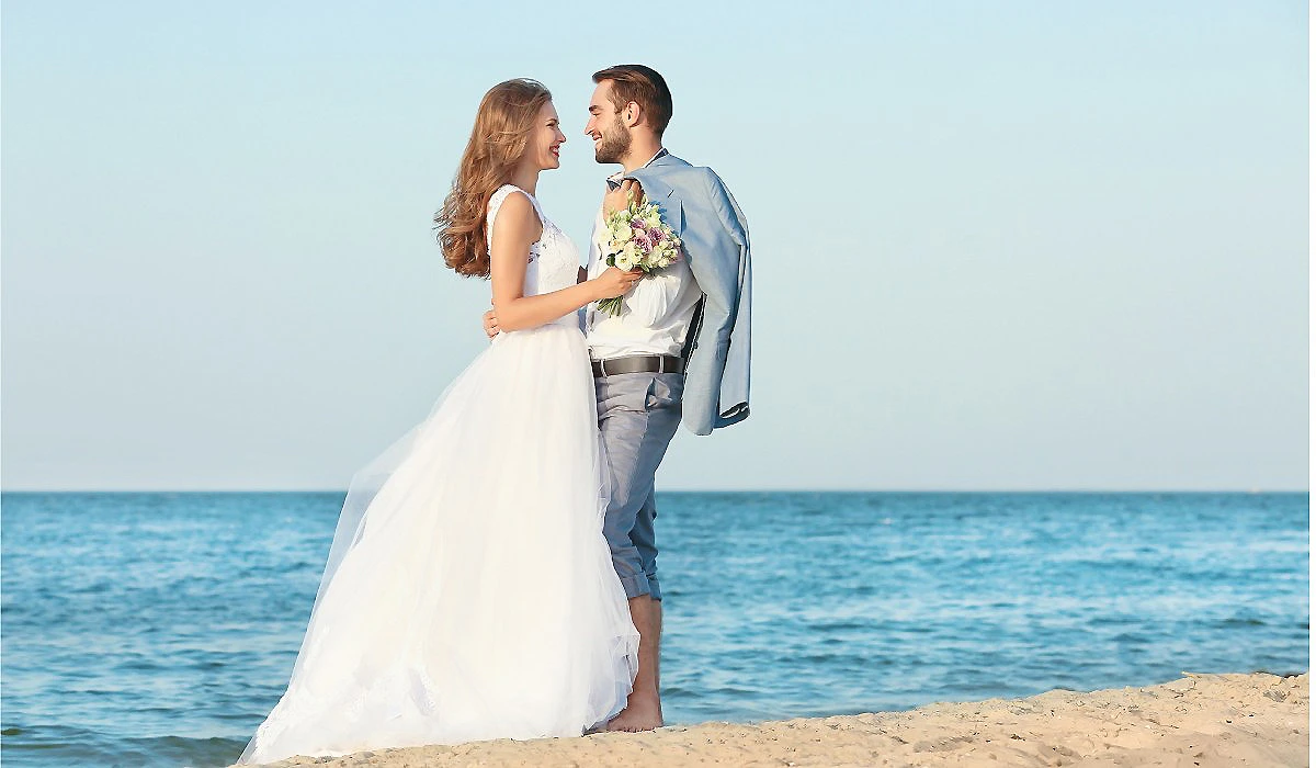 Pareja de novios junto al mar, la novia con ramo y vestido blanco.