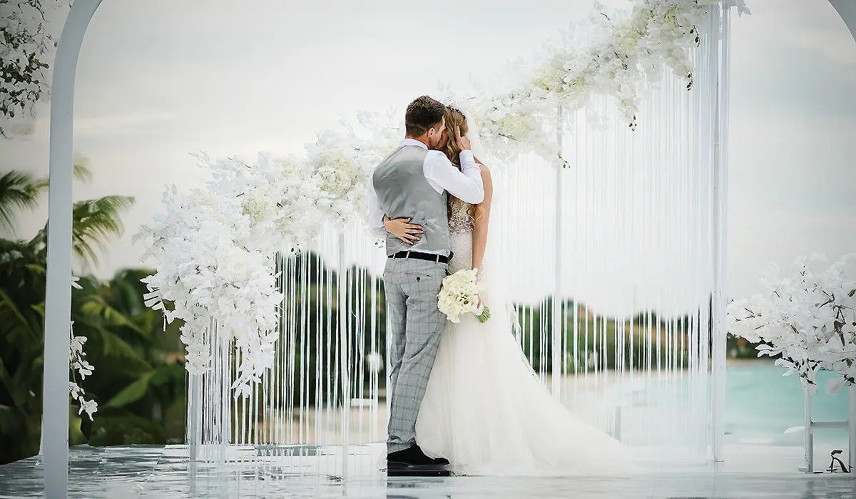 Pareja bailando y abraz&aacute;ndose bajo arco floral blanco en una boda