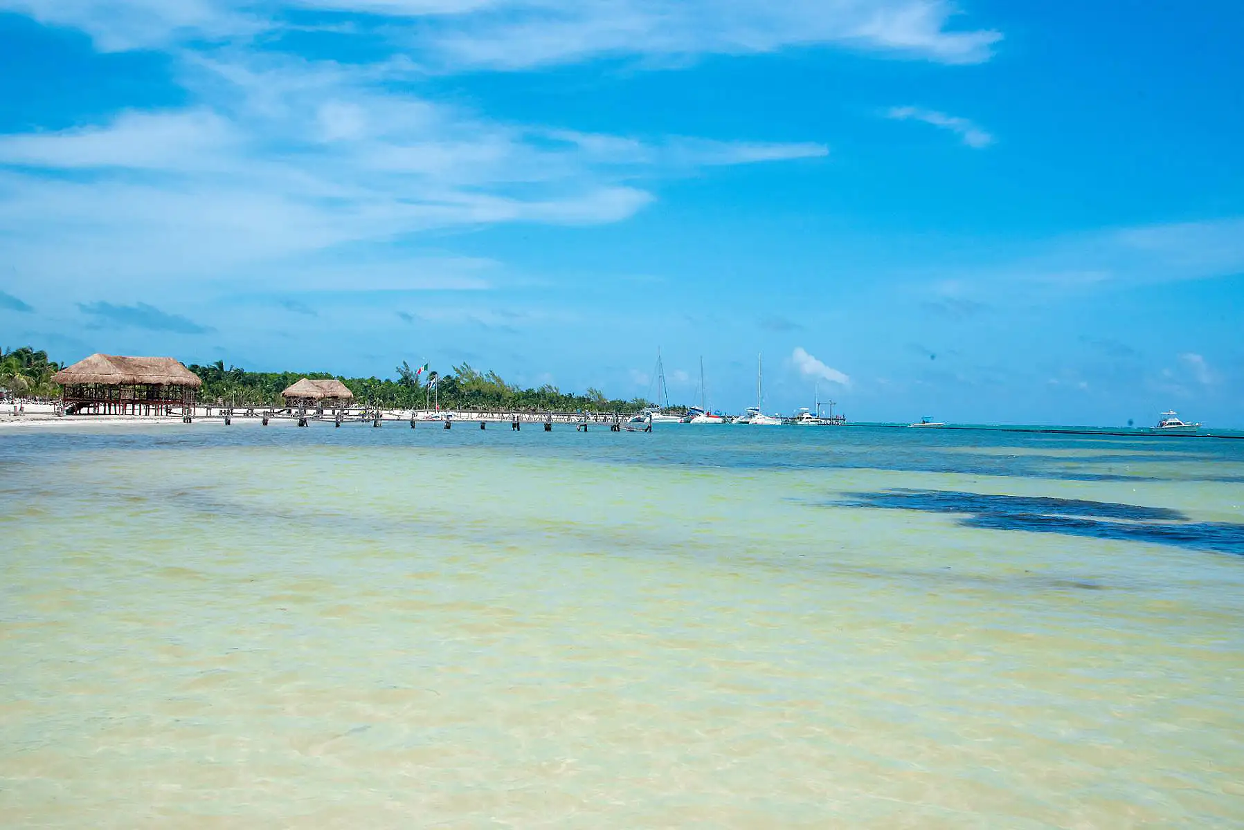 Playa tropical con caba&ntilde;as de paja, agua turquesa y veleros al fondo
