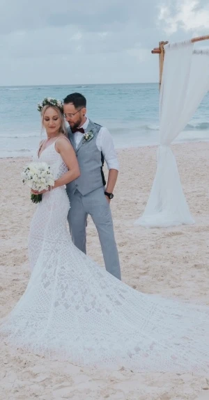Novios en la playa con vestido blanco y ramo, junto al mar.