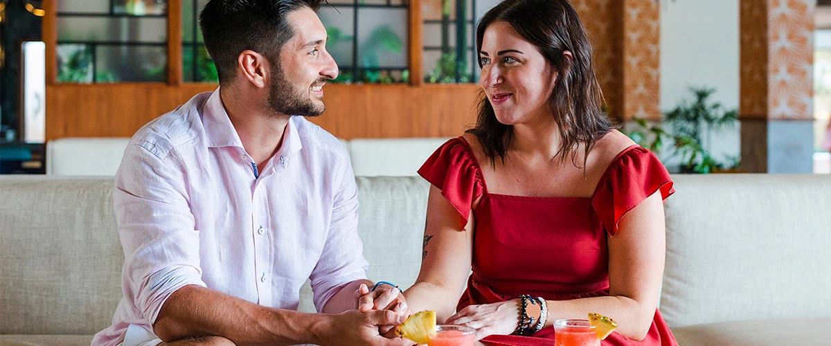 Pareja conversando y sonriendo en un sof&aacute;, con bebidas y sand&iacute;a.