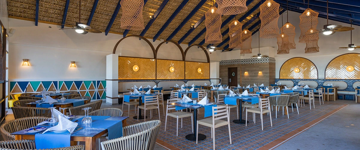 Comedor con mesas azules, sillas de mimbre y l&aacute;mparas colgantes en un hotel.