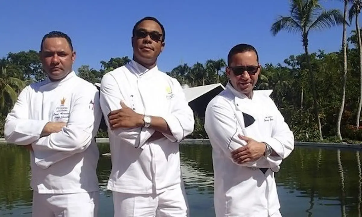 Tres chefs con trajes blancos y gafas, junto a un estanque con palmeras.