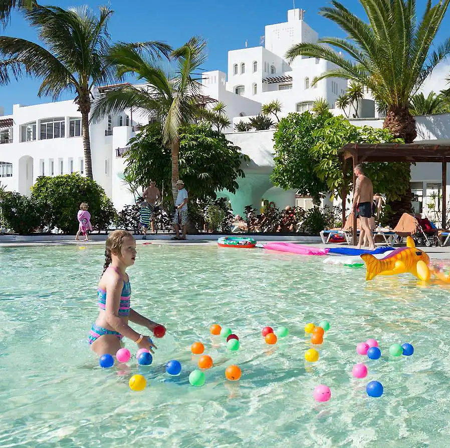 Ni&ntilde;a jugando en la piscina con bolas de colores y palmeras frente a un hotel blanco.
