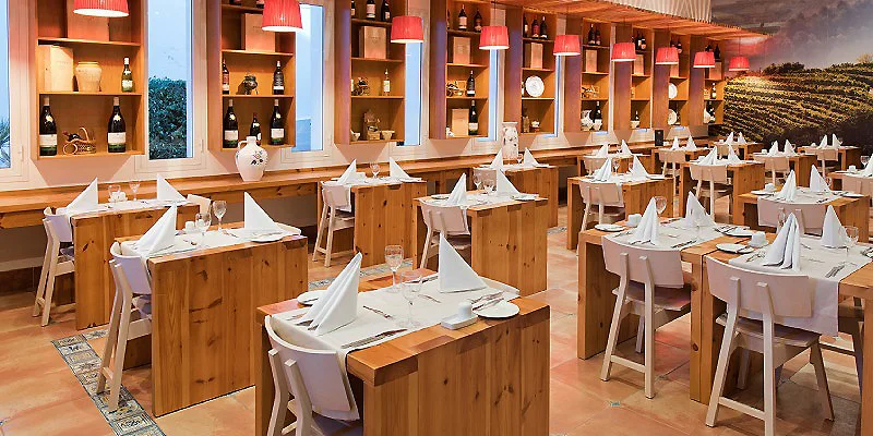 Comedor de restaurante con mesas de madera, sillas blancas y bodega al fondo.