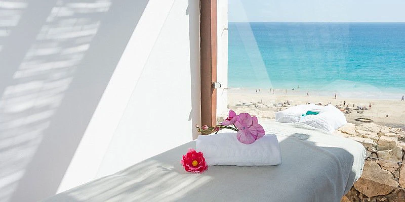 Cama con toallas decoradas y flores frente al mar desde un balc&oacute;n.