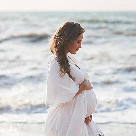Mujer embarazada con vestido blanco, mirando al mar en la playa.