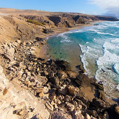 Acantilados &aacute;ridos junto a playa de aguas turquesa y olas.