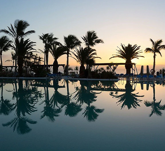 Palmeras y piscina con reflejos al atardecer, cielo al fondo.