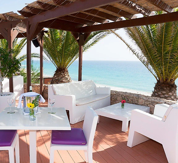 Terraza con mesa blanca, cojines y vista al mar bajo palapas de madera