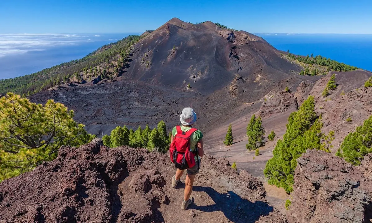 Senderista con mochila sobre volc&aacute;n, junto a monte y oc&eacute;ano al fondo