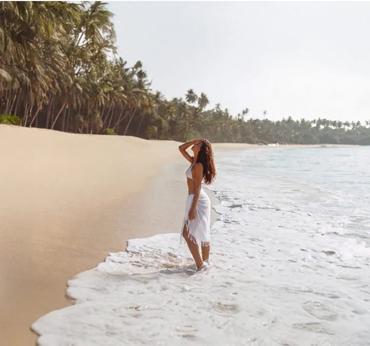 Mujer de pie en la playa, con palmeras y olas al fondo.