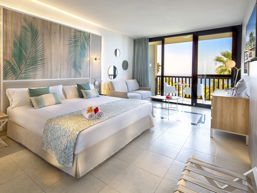 Dormitorio moderno con cama, decoraci&oacute;n tropical y salida a balc&oacute;n con vista al mar