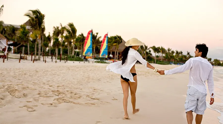 Pareja caminando en la playa con sombrero y ropa blanca, al atardecer