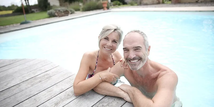 Pareja sonriendo junto a la piscina, relajados en una terraza de madera.