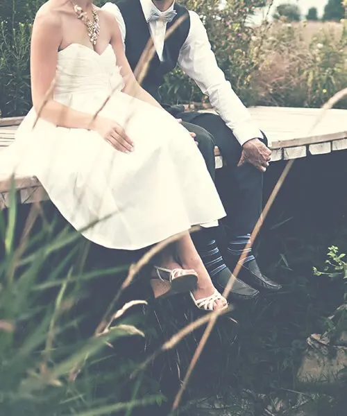 Pareja de novios sentada en un banco, al aire libre entre hierbas y luz.