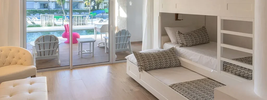 Sala luminosa con camas nido blancas, cojines y salida al exterior con piscina