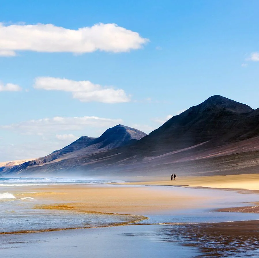 Playa desierta con monta&ntilde;as al fondo y dos personas caminando.