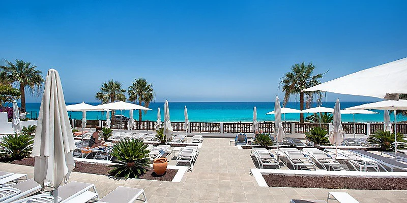 Terraza de hotel frente al mar con tumbonas, sombrillas y palmeras.