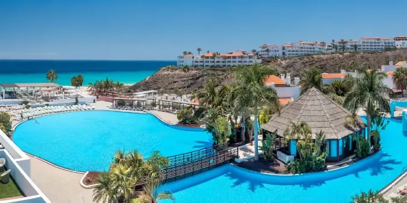 Piscina turquesa de un resort junto al mar, con palapas y palmeras.