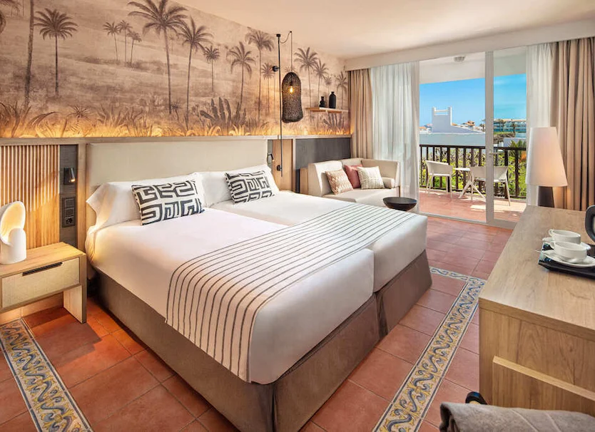 Habitaci&oacute;n de hotel con cama, decoraci&oacute;n tropical y balc&oacute;n con vista al mar.