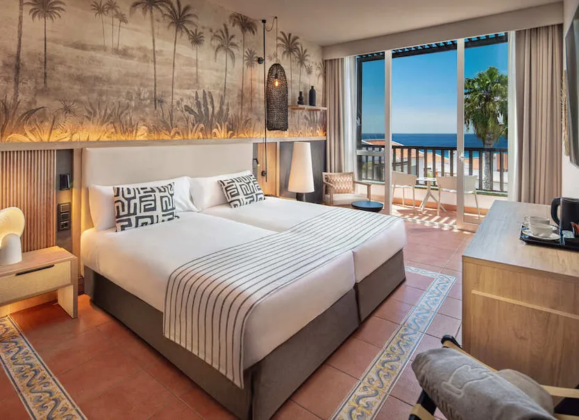 Habitaci&oacute;n de hotel con cama, cortinas y vista al mar desde el balc&oacute;n