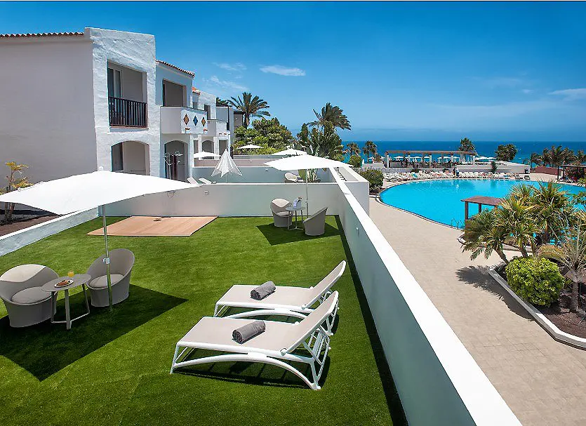 Terraza con c&eacute;sped, tumbonas, piscina azul y vista al mar.