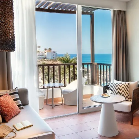 Vista al mar desde un balc&oacute;n con sill&oacute;n y mesa junto a la ventana.