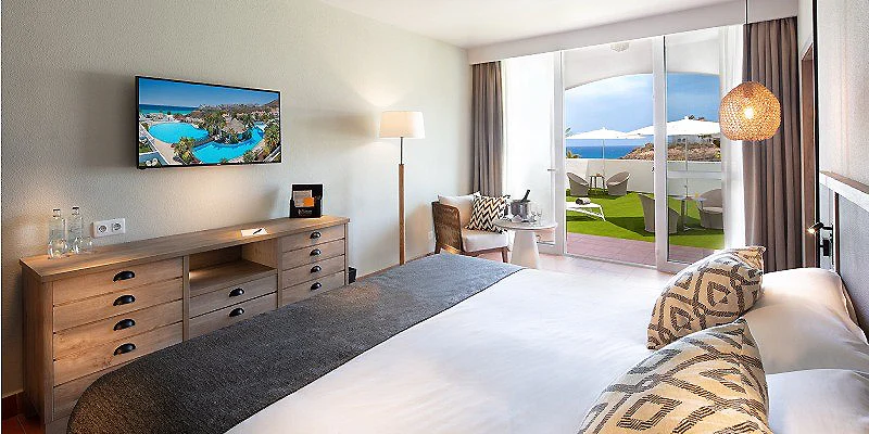 Habitaci&oacute;n moderna con cama, tocador y balc&oacute;n con vista al mar y jard&iacute;n.