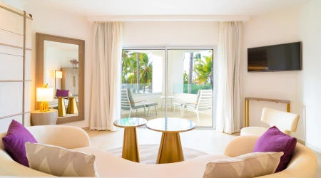 Sala de estar luminosa con sillones, mesa y vista a palmeras por la ventana.