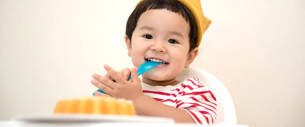 Beb&eacute; sonriente con gorro amarillo, comiendo con cuchara azul y mamila.