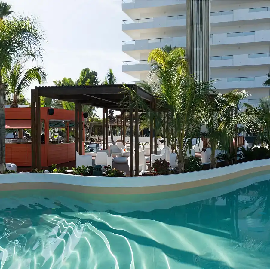 Piscina con agua y plantas tropicales frente a un hotel moderno de varios pisos
