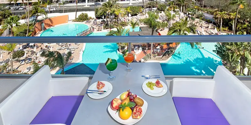 Mesa con frutas y copas frente a piscina azul y palmeras en el hotel
