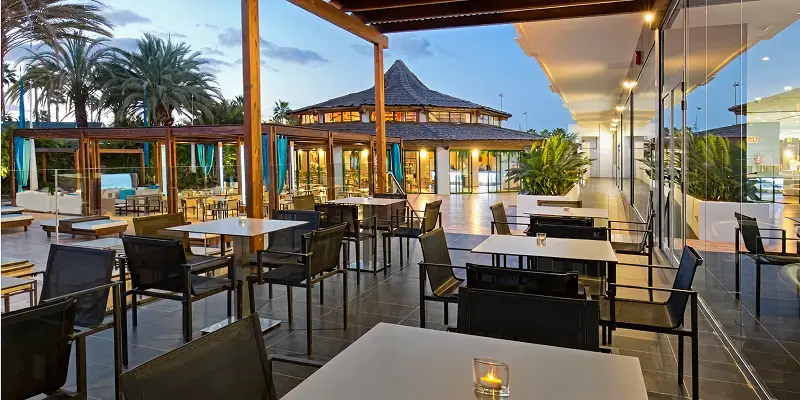 Terraza de hotel con mesas y sillas, palmeras y luz al atardecer.
