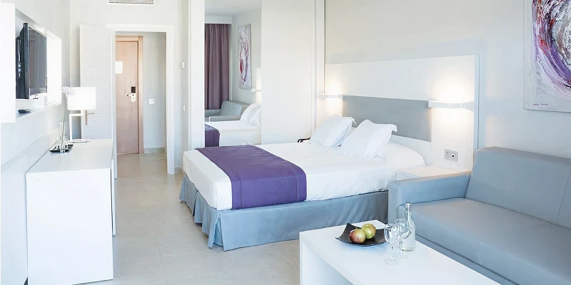 Habitaci&oacute;n de hotel moderna con cama doble, sof&aacute; y mesa con frutas y copas.