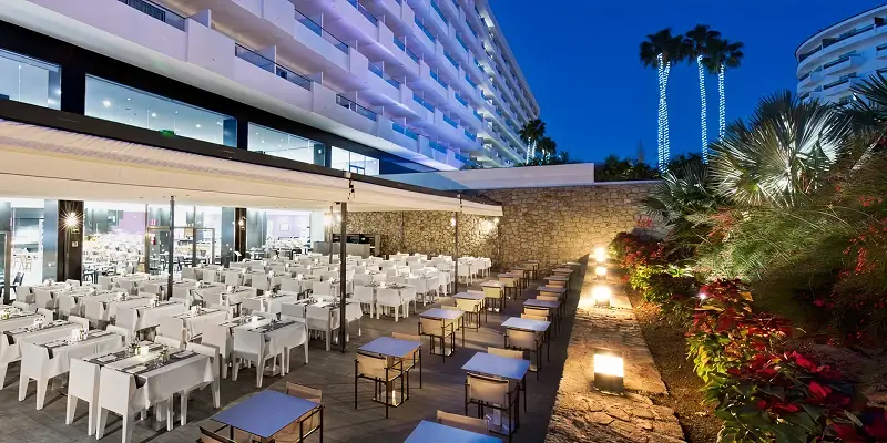 Terraza de hotel con mesas blancas y palmeras iluminadas de noche