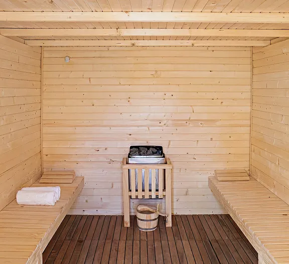 Interior de sauna de madera, con bancos, balde y estufa al centro.