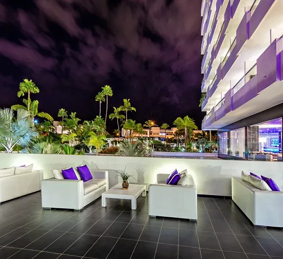 Terraza nocturna moderna con sof&aacute;s blancos y jard&iacute;n con palmeras iluminadas