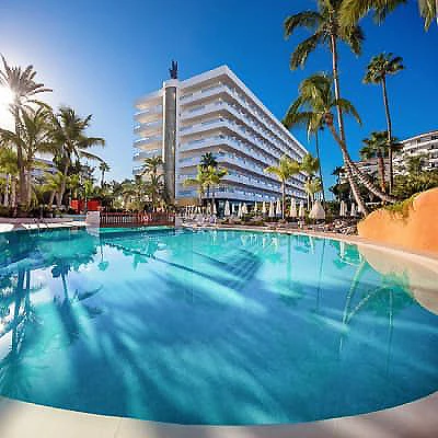 Piscina con palmeras y hotel blanco junto al mar, cielo azul tropical.