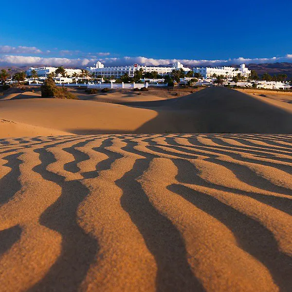 Hotel blanco y dunas con sombras onduladas bajo un cielo azul