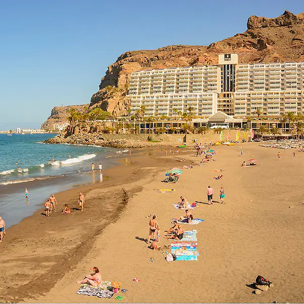 Playa de arena con ba&ntilde;istas y hotel frente a acantilado en la costa.